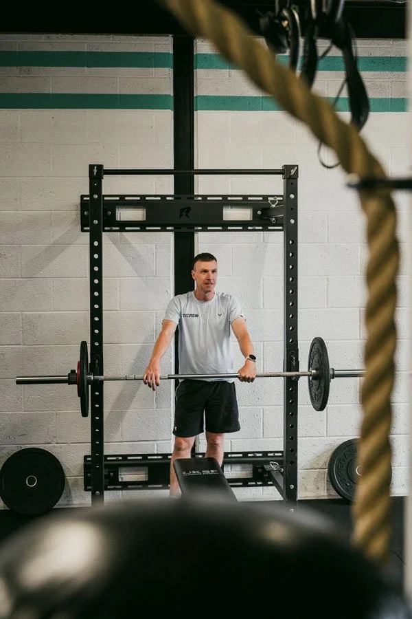Adam holding a loaded barbell inside a gym squat rack