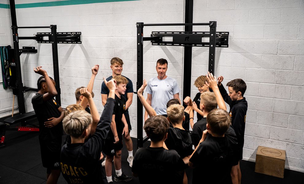 Youth sports team gathered with coaches Adam & Felix in a gym near weight racks, raising their hands during a group activity.