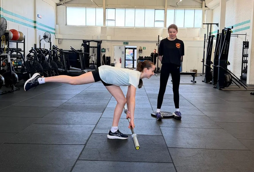 Girl performing a single-leg deadlift with a barbell in a gym as a another observes.