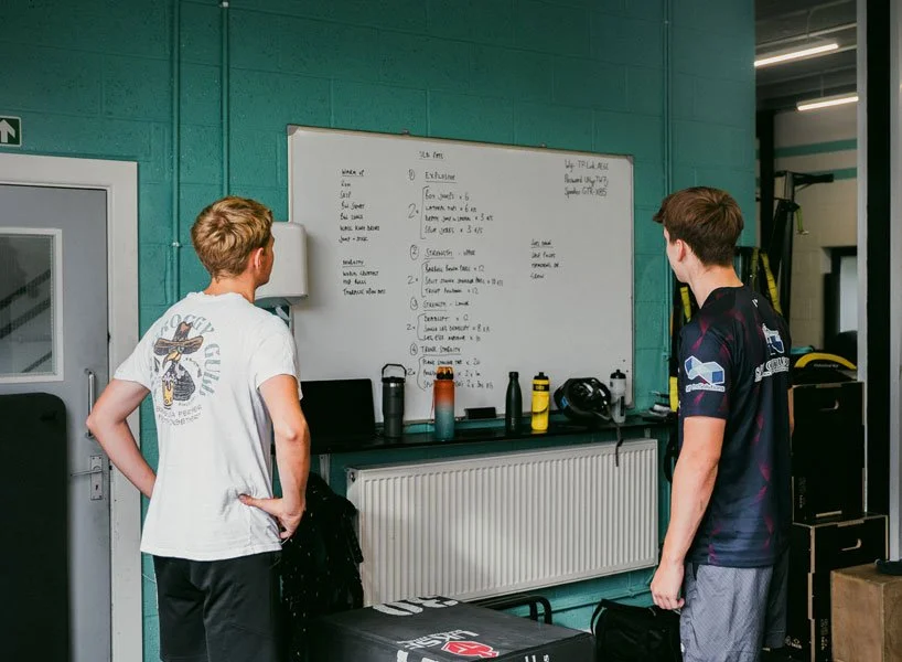 Two athletes reviewing a whiteboard training plan in a teal-walled locker room