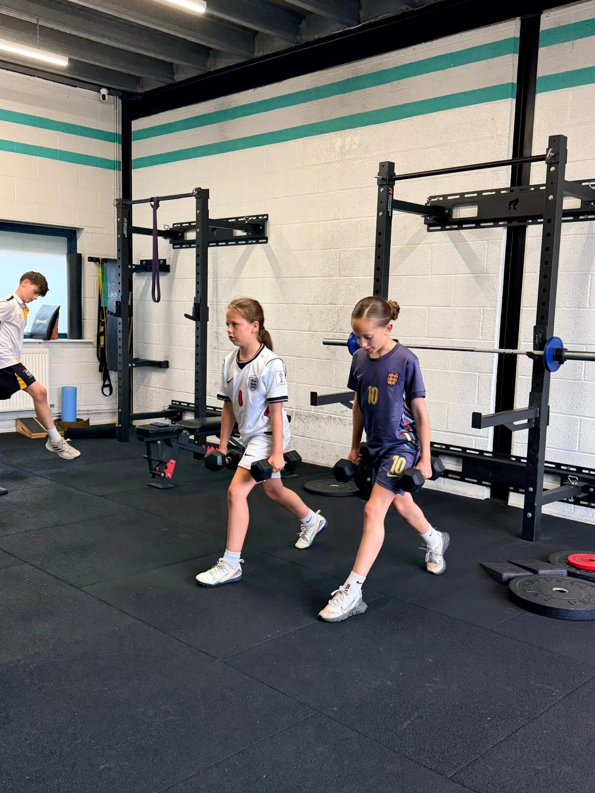 Two young female soccer players perform walking lunges with dumbbells in a gym, with squat racks and weight plates behind them.