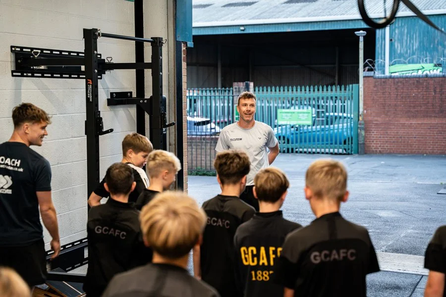 Youth soccer players listening to a coach Adam Lloyd inside a gym near an open warehouse door