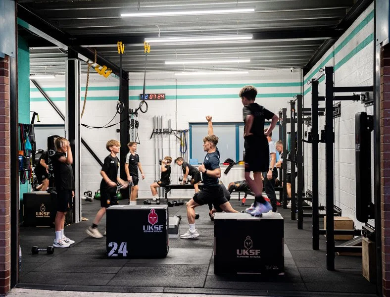 Youth athletes training in a functional fitness gym with plyometric boxes, squat racks, kettlebells, rings, and ropes.