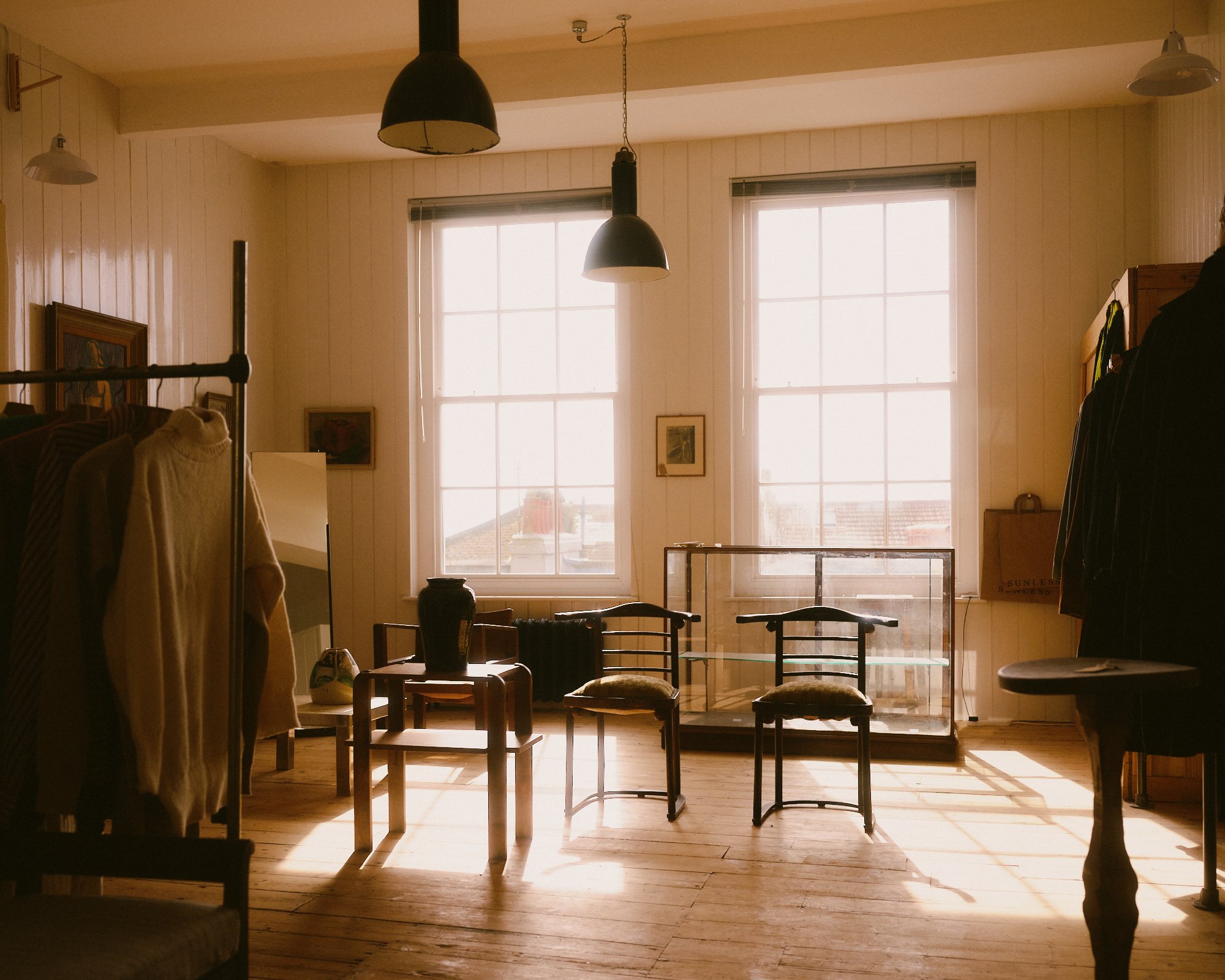 A photograph of a historic shop interior with a minimal feel. Timeless vintage and antique clothing is displayed throughout interspersed with modernist furniture.