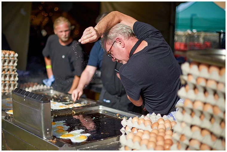 Chef preparing breakfast eggs on a large griddle at a festival crew catering kitchen, surrounded by stacked egg trays ready for service