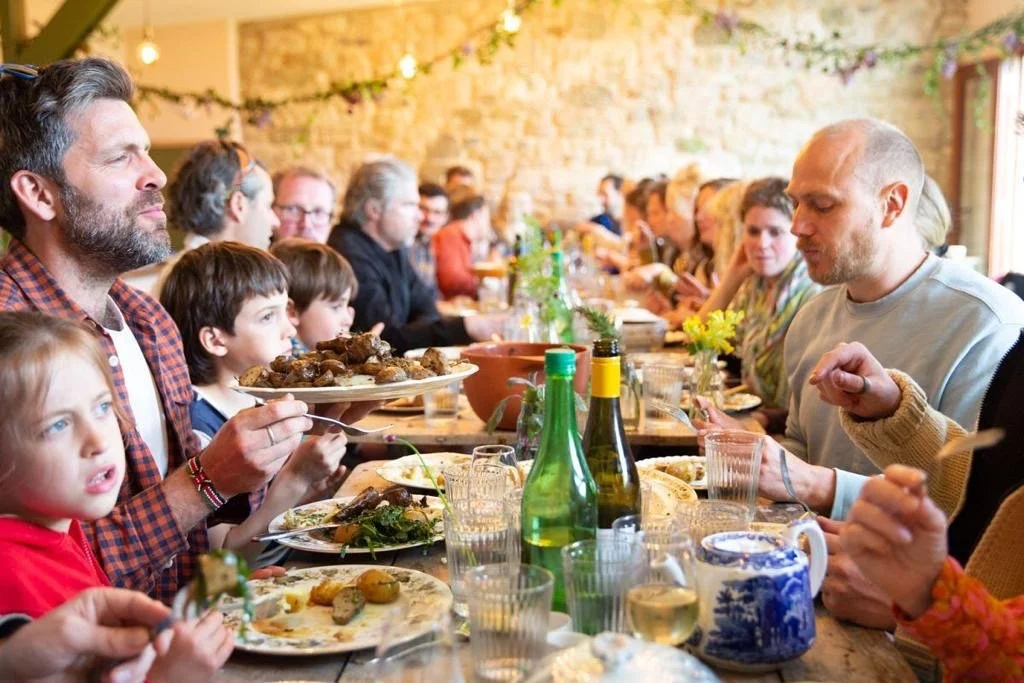 Festival guests of all ages gathered around a long communal feasting table, sharing family-style platters of food lit by warm string lights against a stone wall backdrop