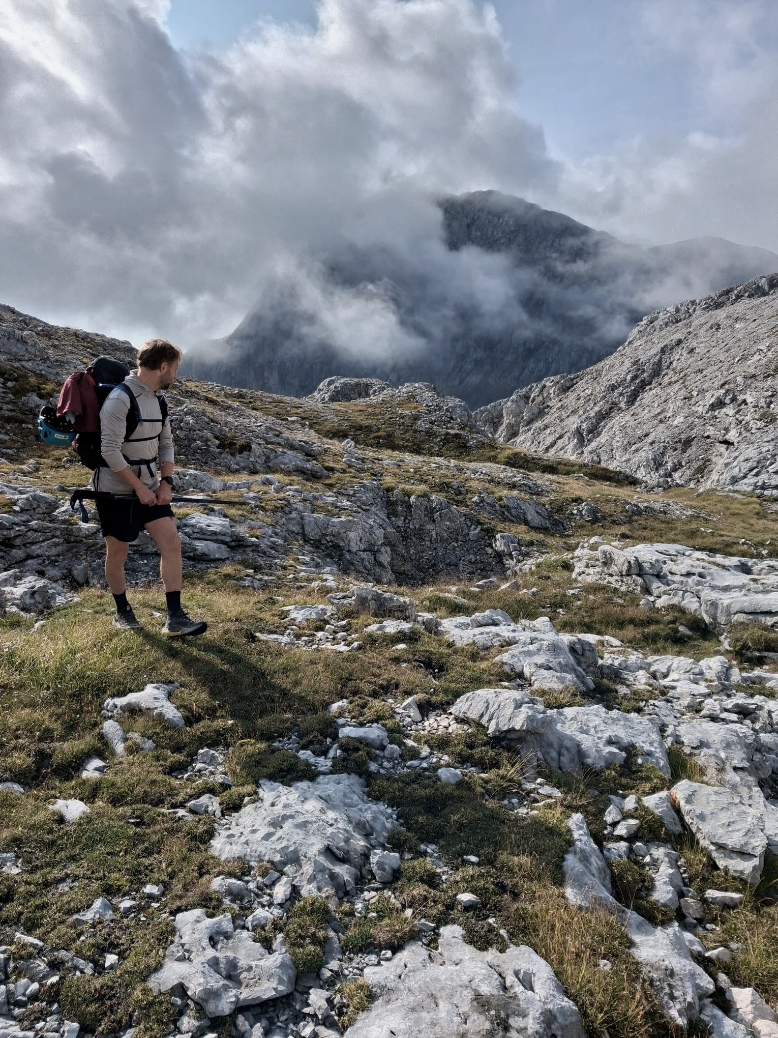 A person hiking on a rocky mountain trail surrounded by large rocks and grassy patches, with a mountain peak shrouded in clouds in the background.