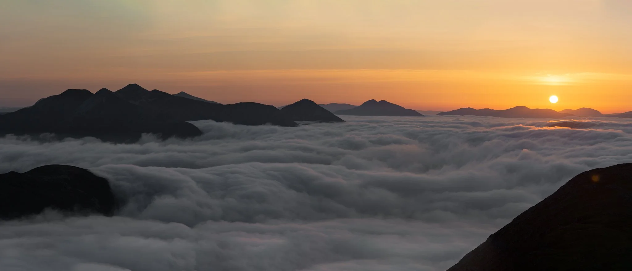Sunset over a mountain range with a thick layer of clouds below and a partially visible sun on the horizon.