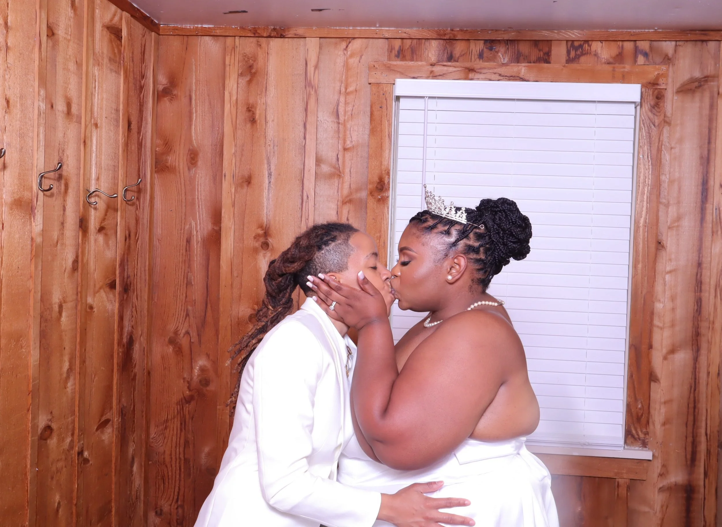 Two women, one dressed in a white suit and the other in a wedding gown with a tiara, share a kiss in a wooden room.