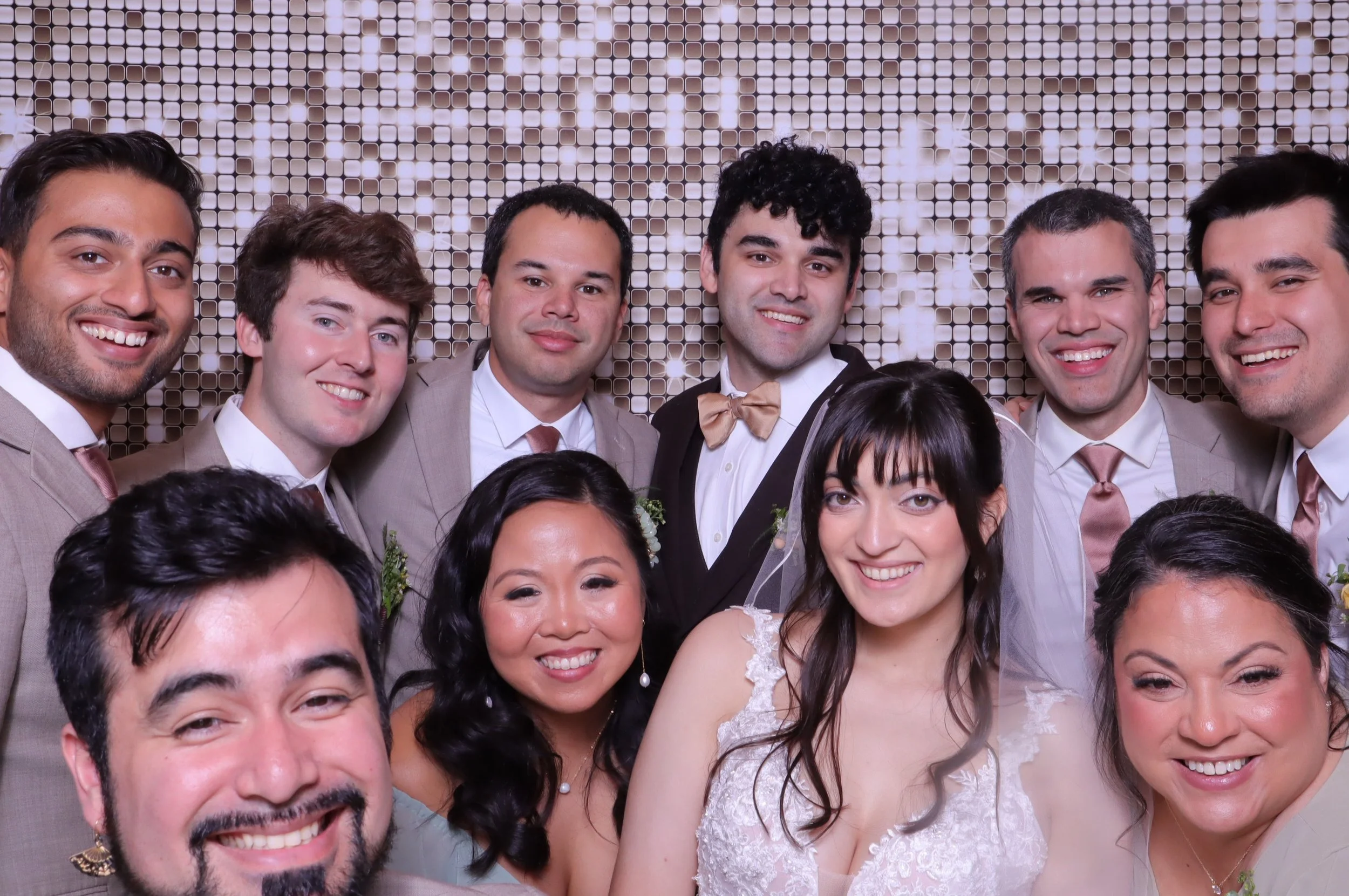 Wedding party dressed in wedding attire smiling for photo booth pictures in front of a gold sparkly backdrop.