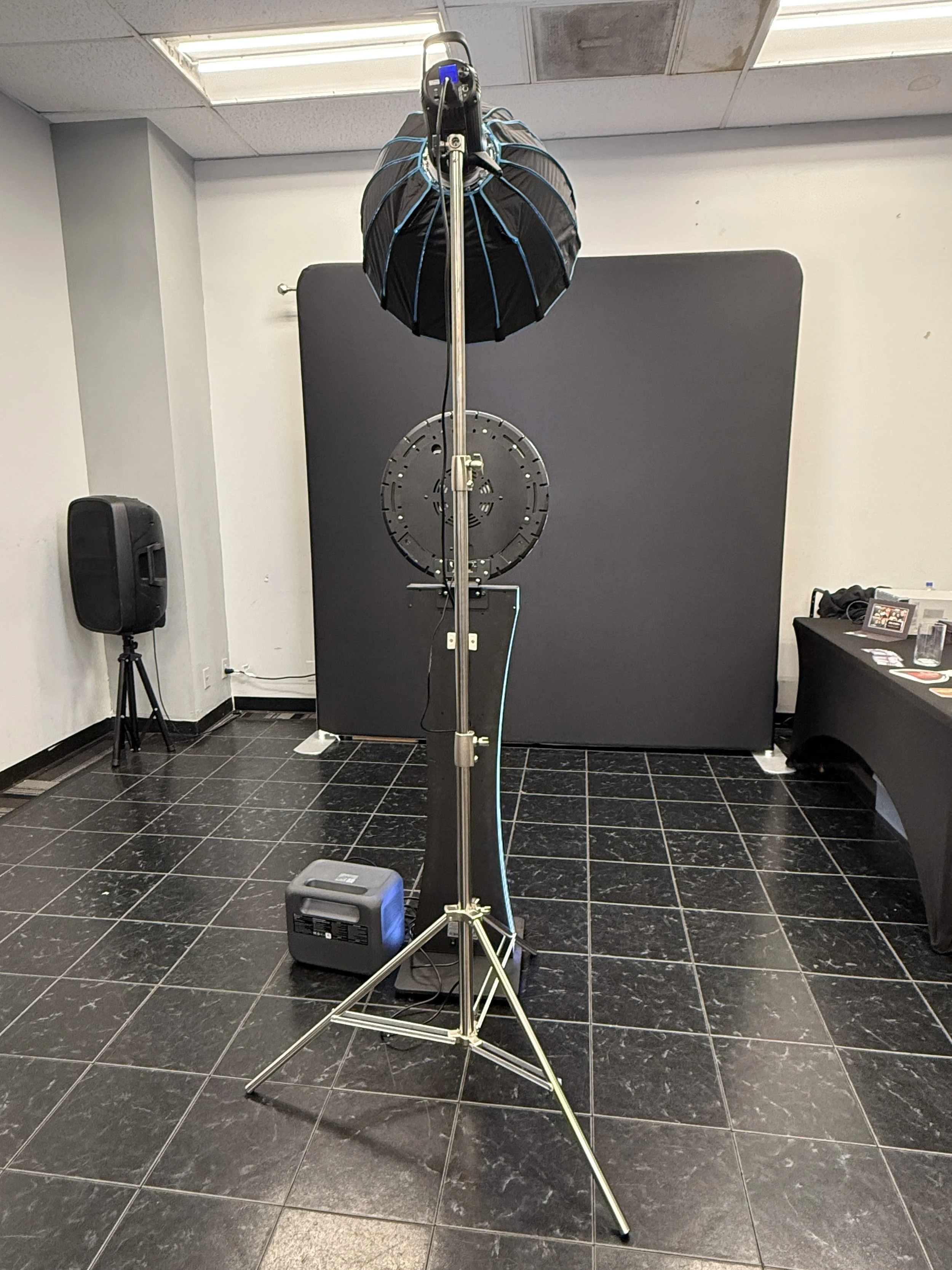 Photo of professional photography lighting equipment with a large umbrella reflector, light stand, and a softbox, set up on a black tiled floor in an indoor setting with a black backdrop.