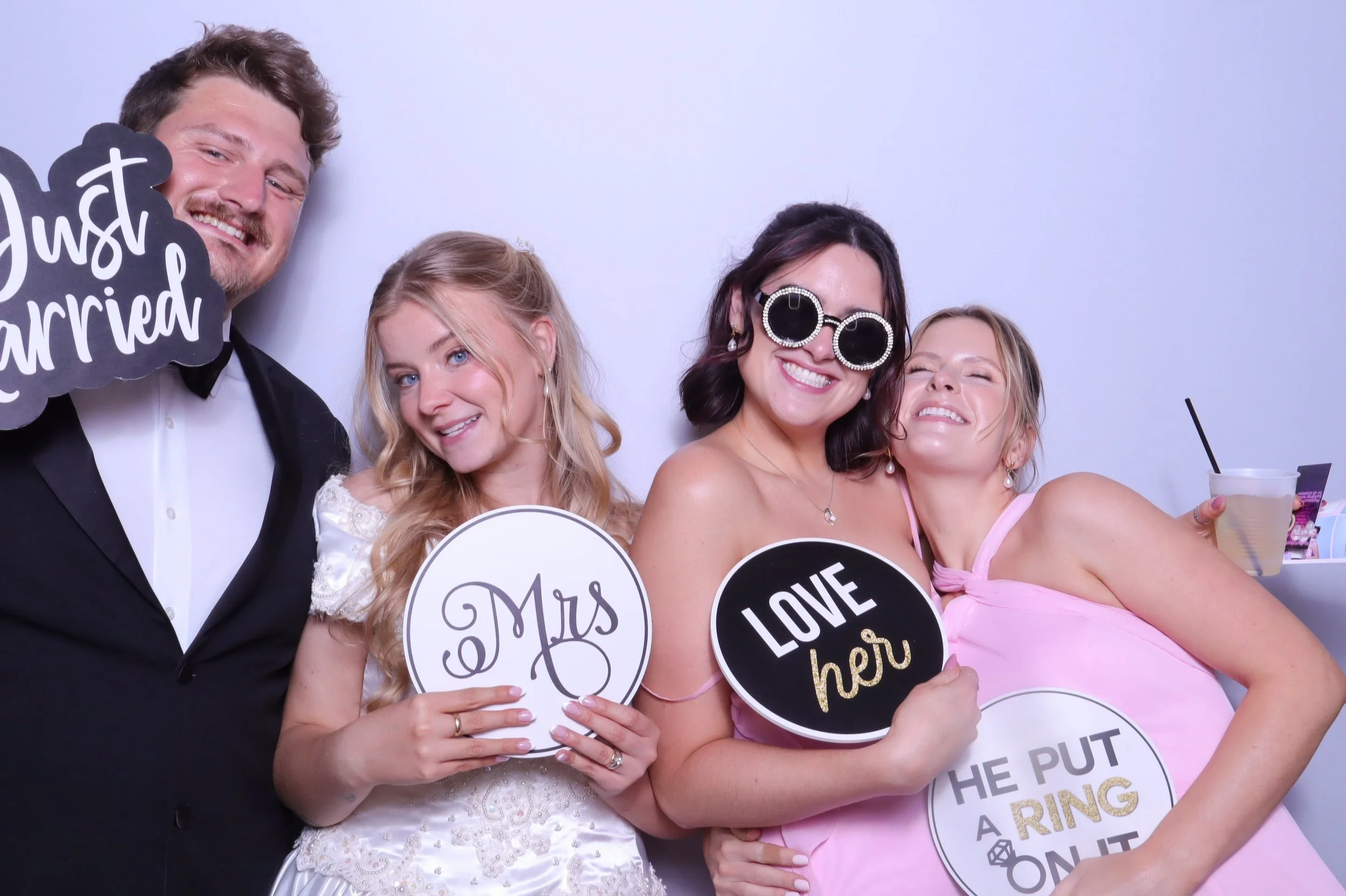 Four friends celebrating a wedding, holding signs that say "Just Married," "Mrs," "Love her," and "He put a ring on it," smiling and dressed in formal attire.