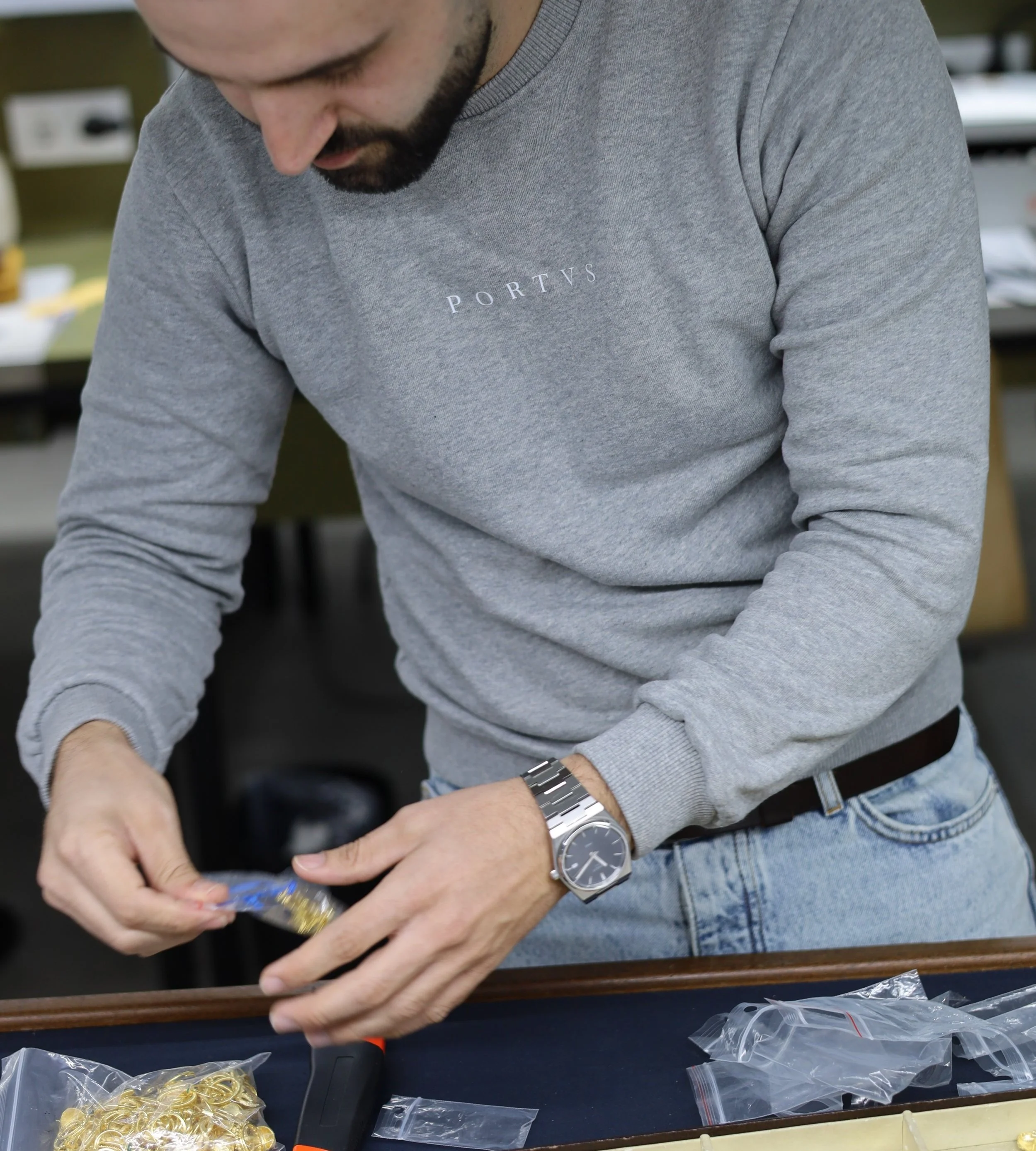 Homem organizando joias ou peças douradas em sacos plásticos, usando um moletom cinza e relógio de pulso.