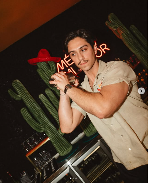 Homem preparando coquetel atrás de um bar com cactos e letreiro neon ao fundo.