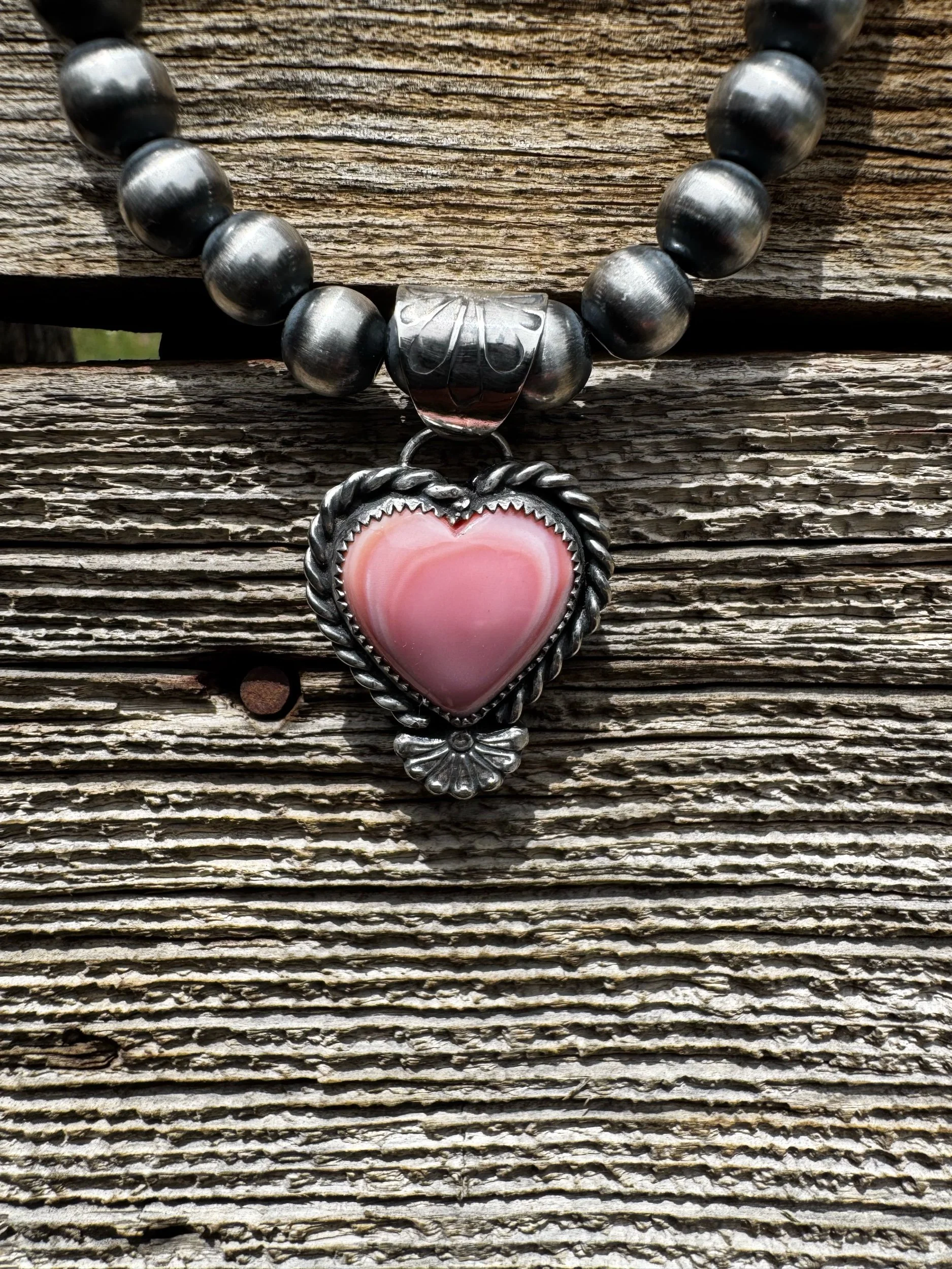 A necklace with dark beads and a heart-shaped pink stone pendant hanging on a weathered wooden surface.