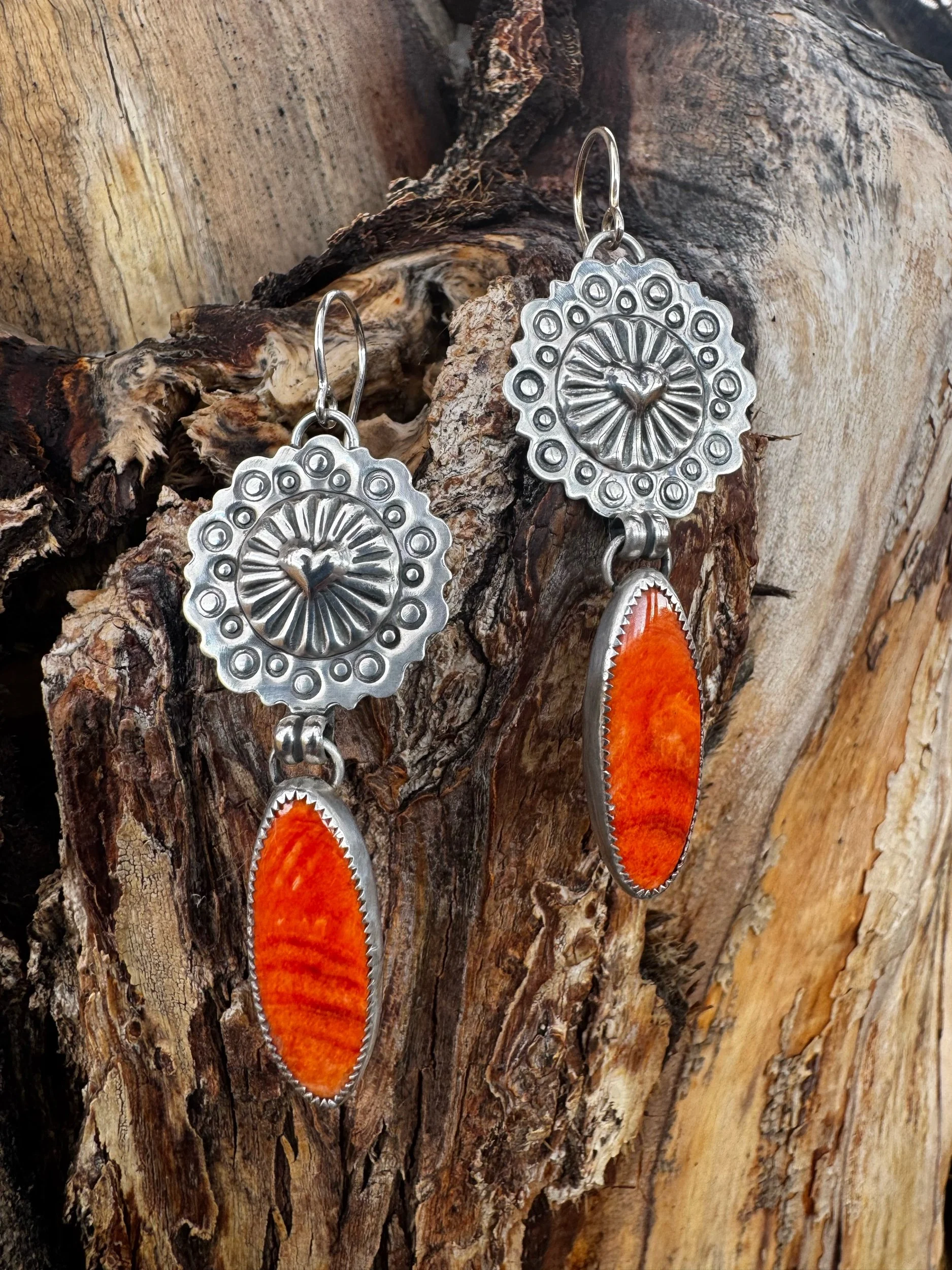 Silver earrings with circular floral design featuring orange oval stones, hanging from a piece of weathered wood.