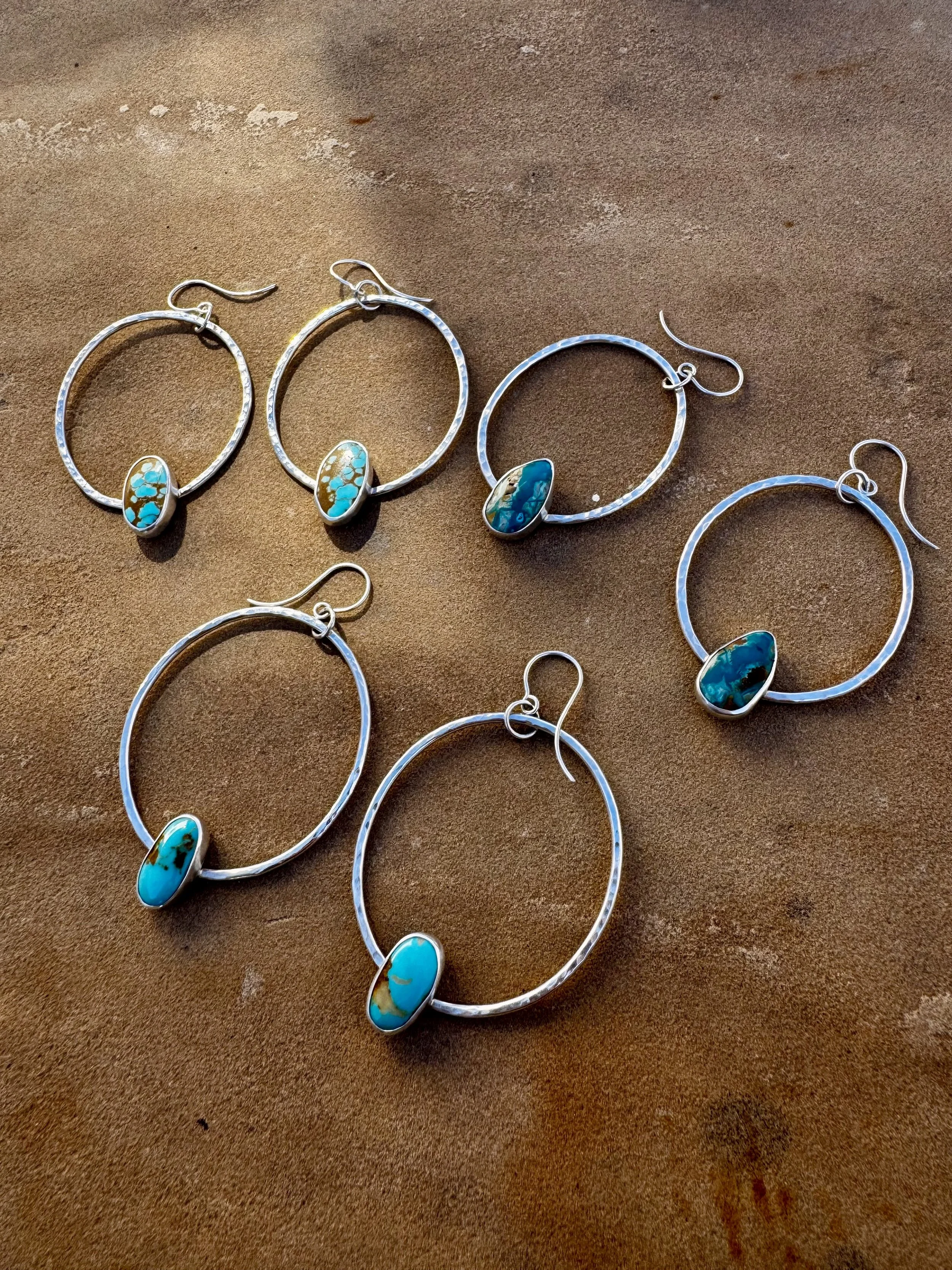 Six pairs of silver hoop earrings with turquoise stones on a brown surface.