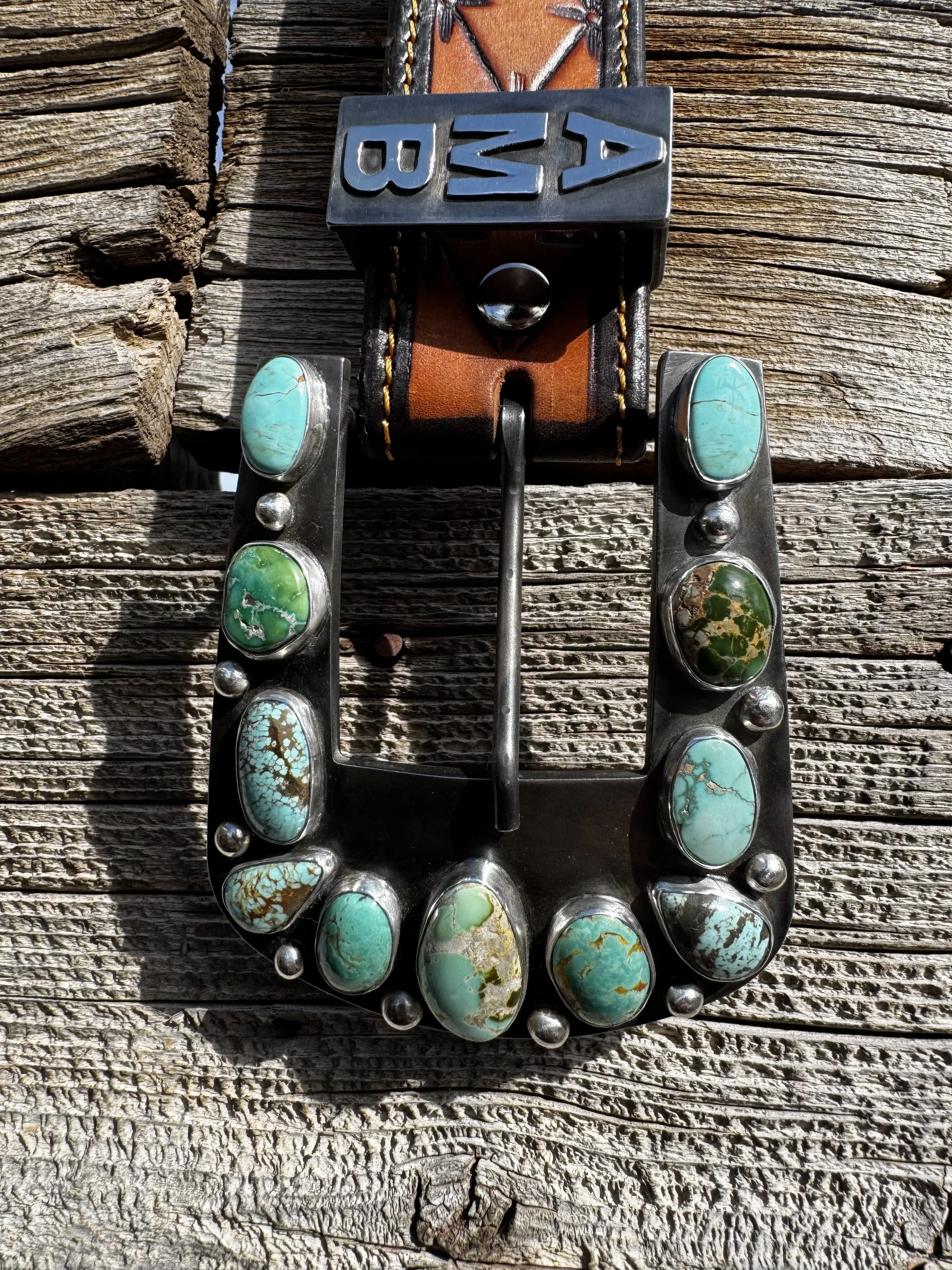Western-style belt buckle with turquoise stones, attached to a leather belt, mounted on a weathered wooden surface.