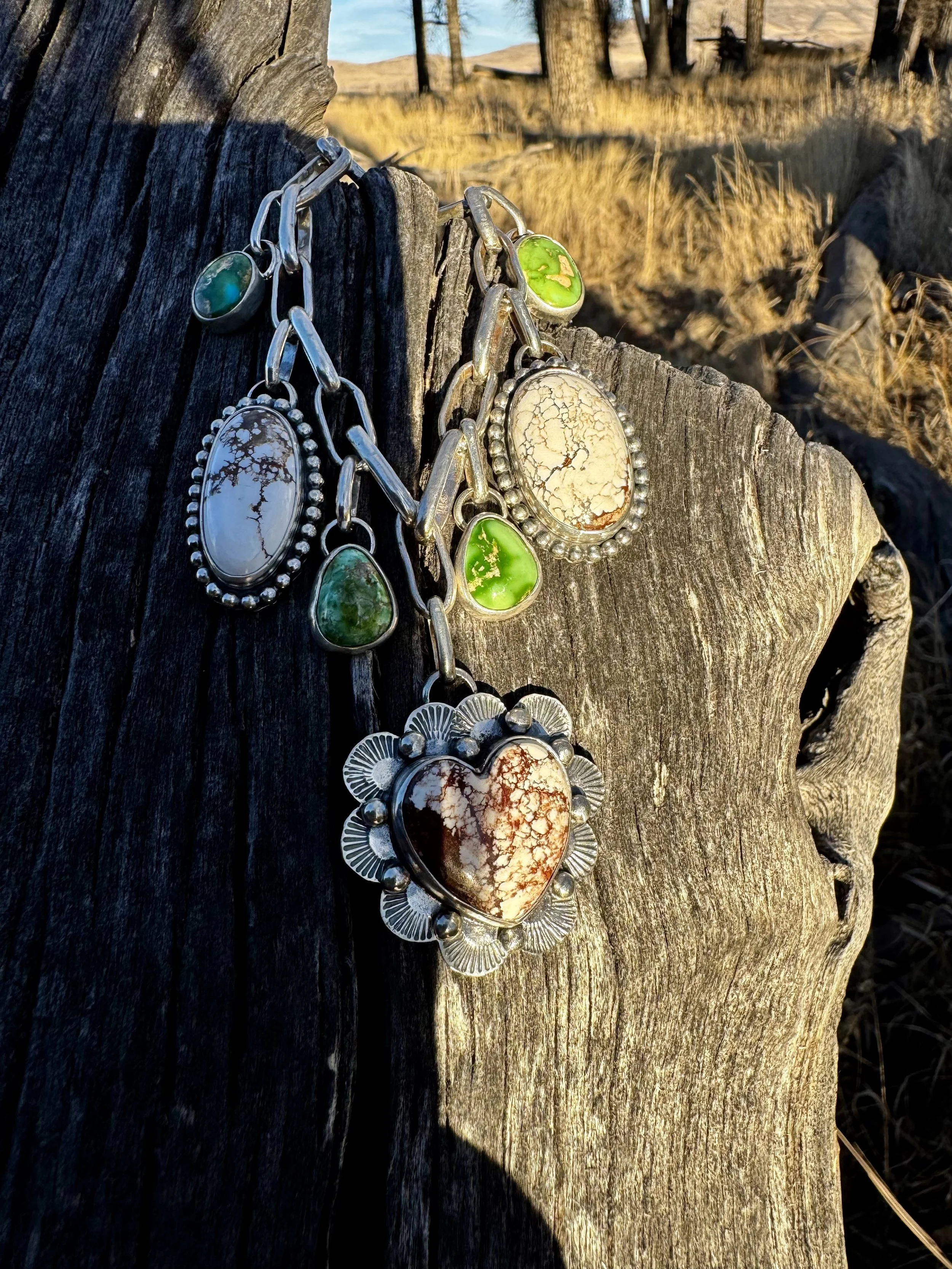Silver jewelry with turquoise and green stones hanging on a weathered wooden post in a desert landscape.