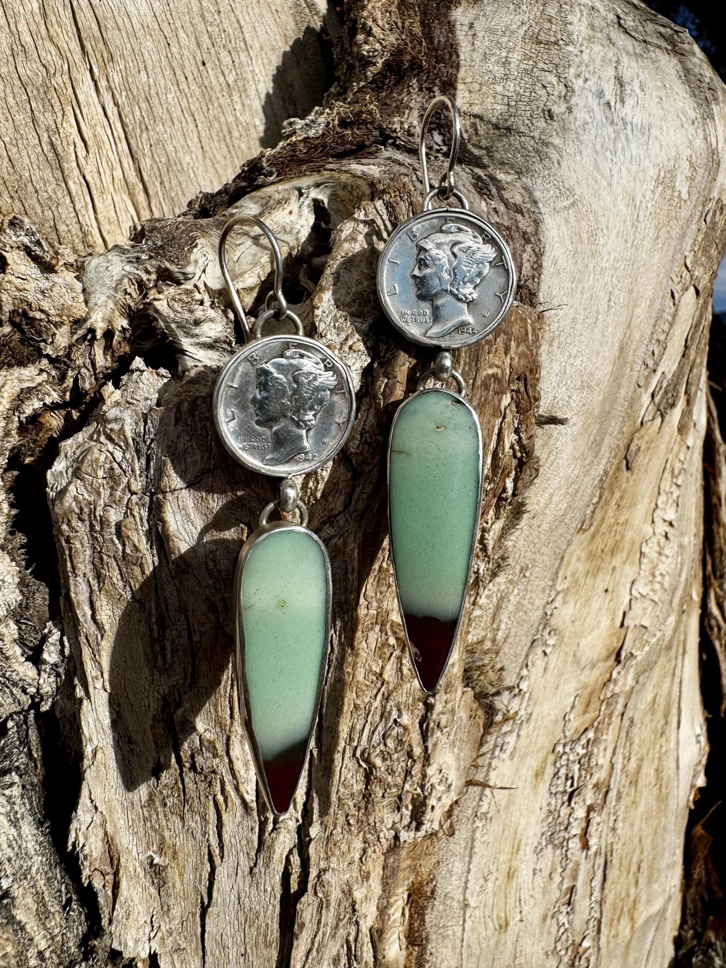 Earrings featuring a coin replica with a woman's profile and a long, pointed, multicolored stone pendant with shades of green and brown, hanging on a textured piece of wood.