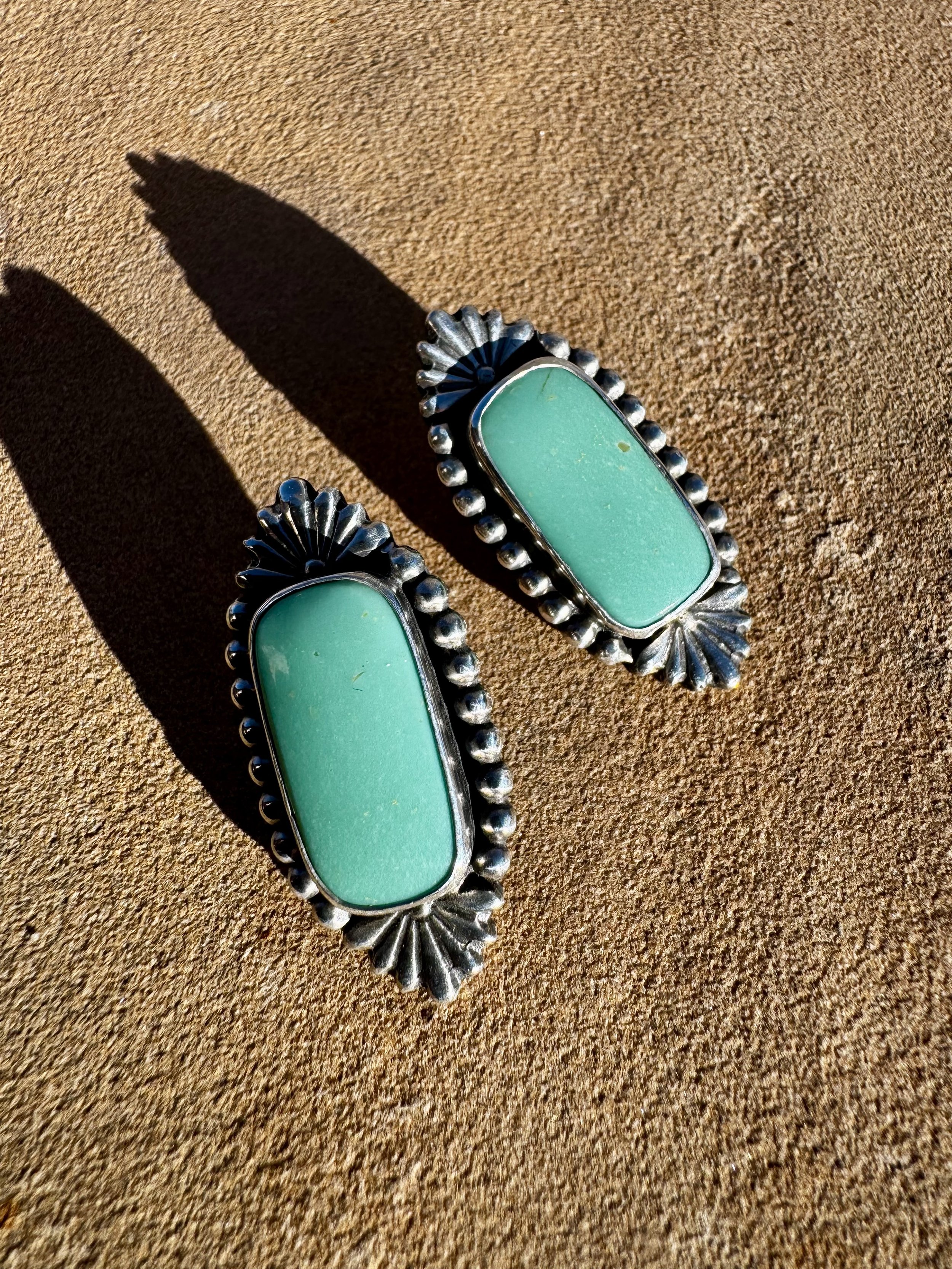 Pair of turquoise and silver Native American-style earrings on sand, casting shadows.