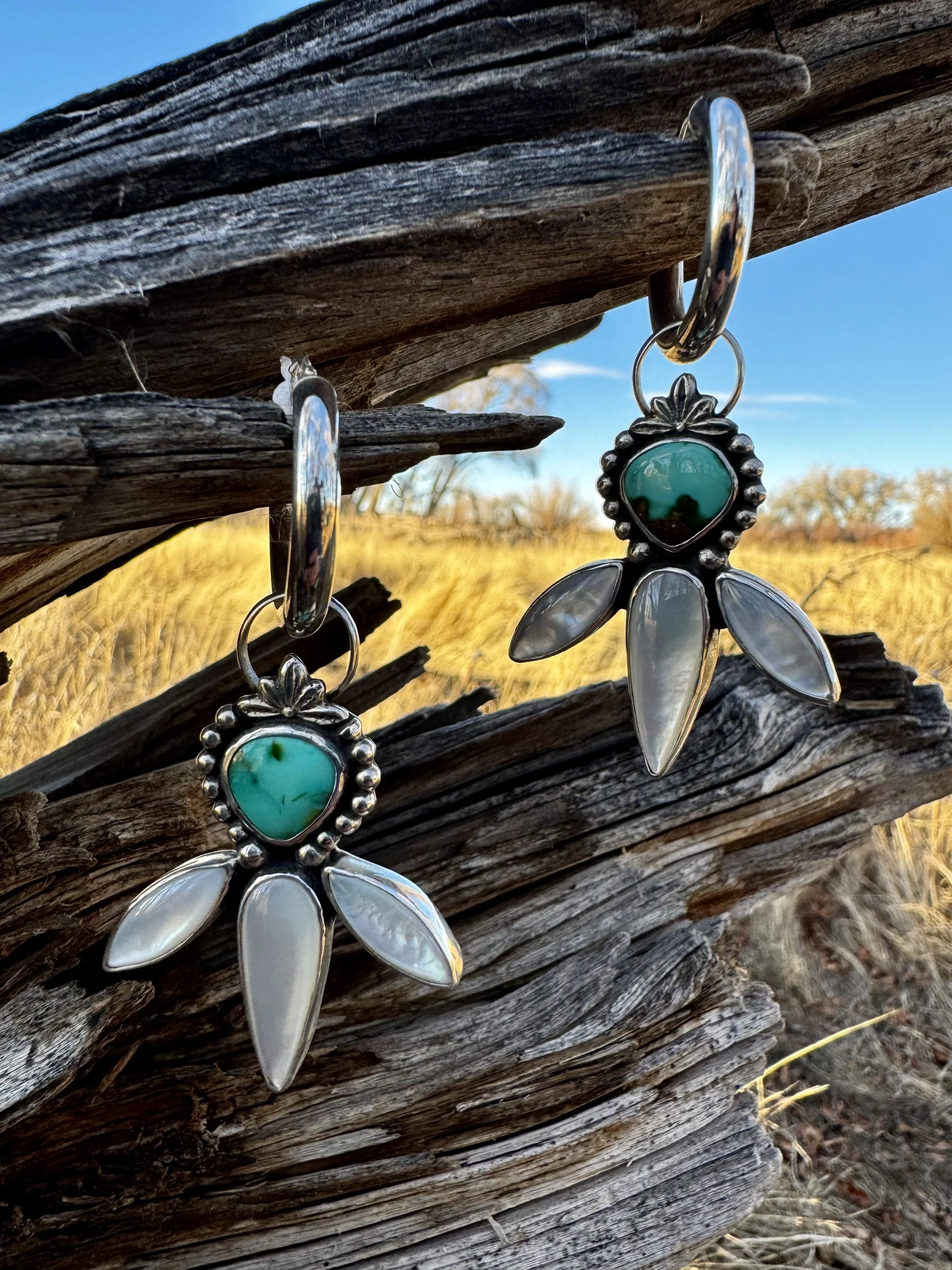 Pair of silver earrings with turquoise and shell-like stones, hanging on a weathered wooden branch outdoors with a grassy field and blue sky in the background.
