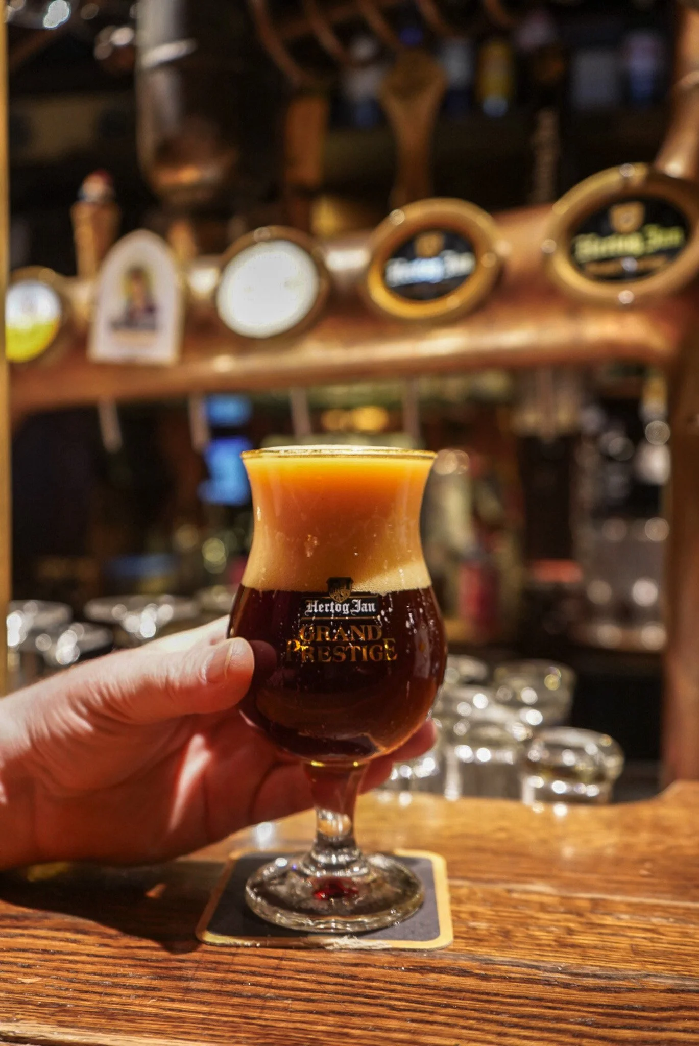 A hand holding a tulip-shaped glass of dark beer with a foamy head, labeled 'Hertog Jan Grand Prestige', on a wooden bar counter. The background shows a bar with taps and blurred liquor bottles.