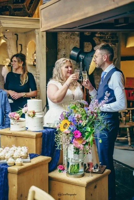 A bride and groom toast with champagne at their wedding reception in a rustic setting, surrounded by wedding cakes and a large floral arrangement.