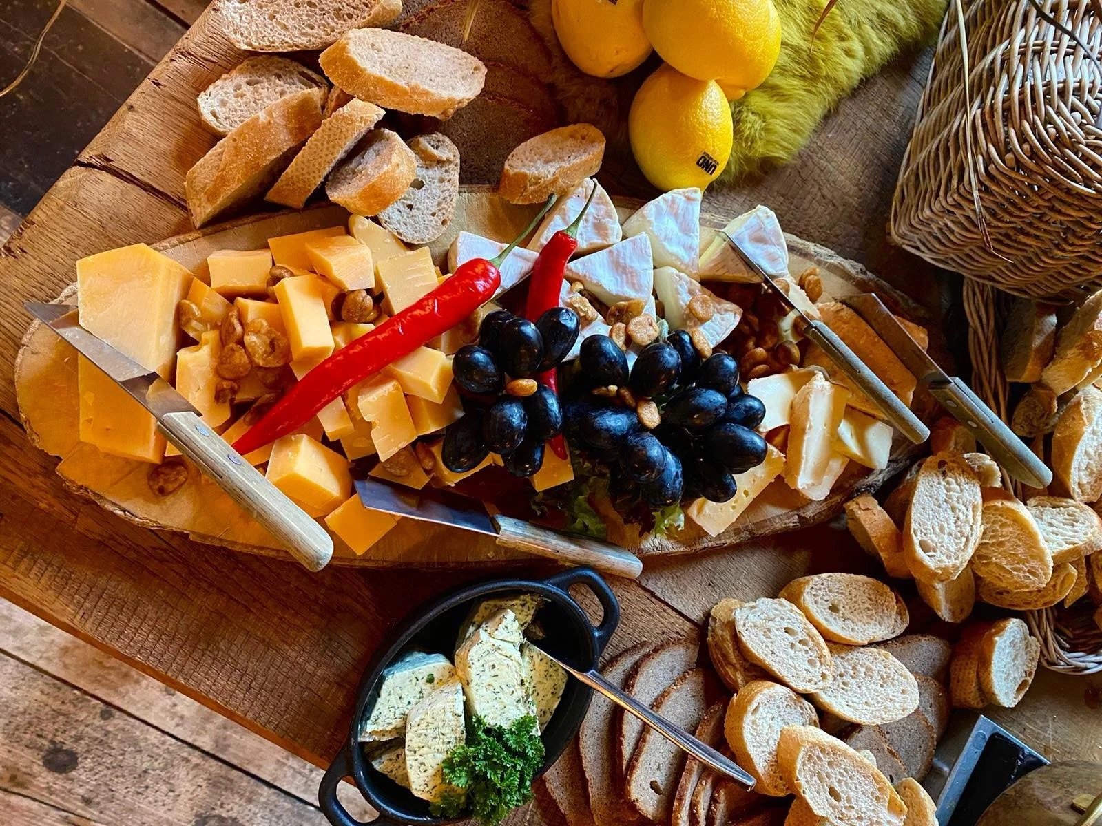 A cheese and fruit platter with various cheeses, black grapes, red chili pepper, and nuts, surrounded by slices of baguette on a wooden serving board.