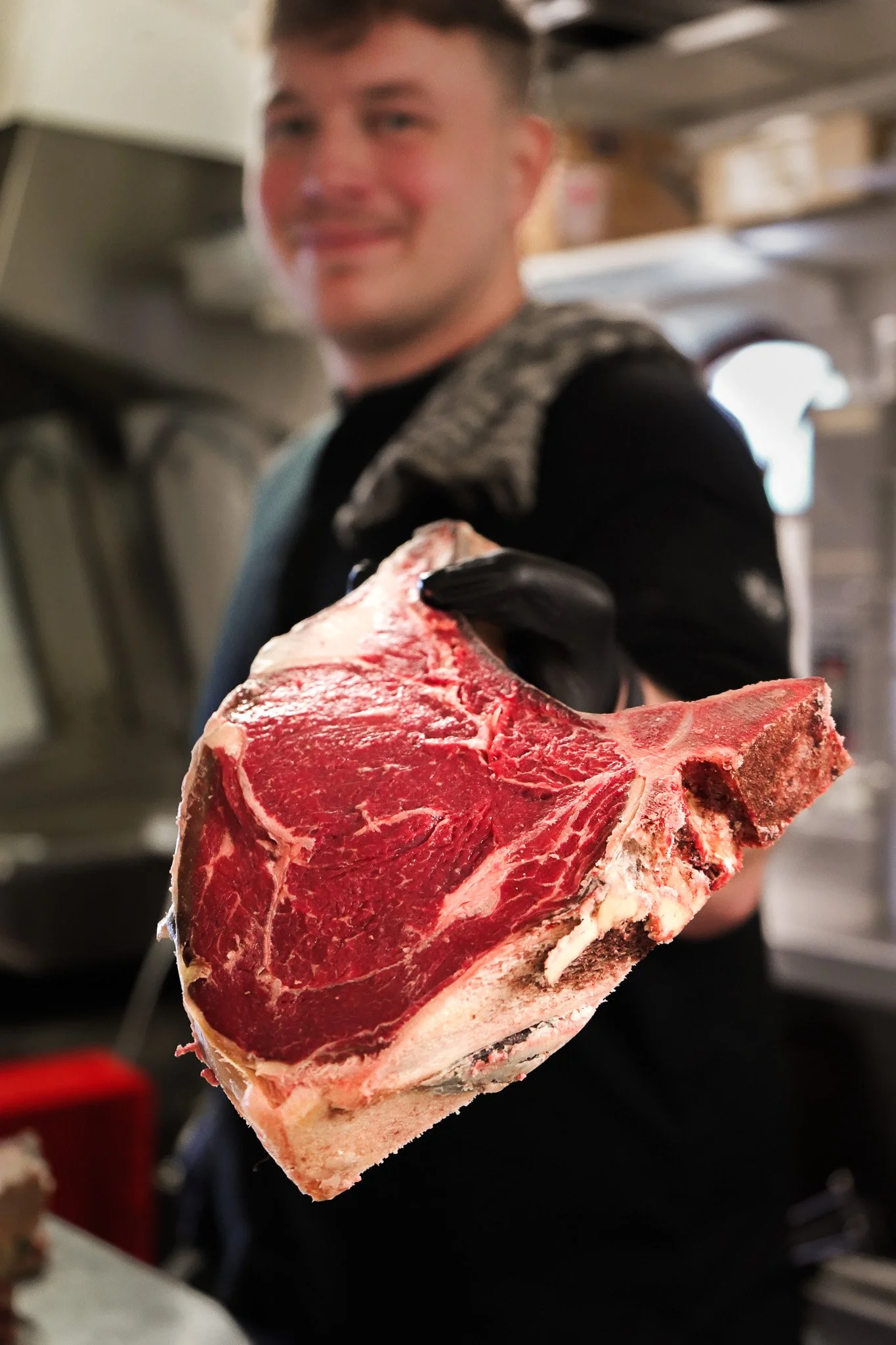 A person holding a large raw T-bone steak towards the camera, with a blurred background of a kitchen or butcher shop.