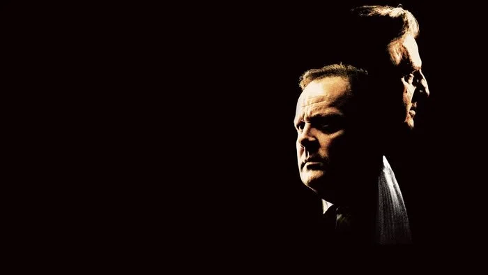 A dark image featuring two men's faces, one in the foreground and one in the background, both looking serious, with dramatic lighting highlighting their features.