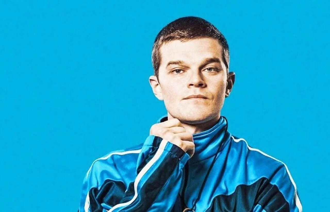 A young man with short brown hair wearing a blue athletic jacket stands against a bright blue background, with his hand resting on his chin, looking thoughtful.