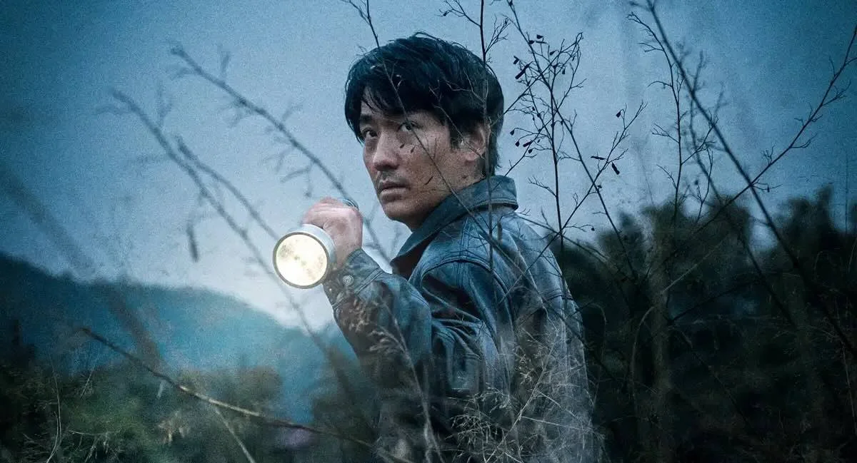 A man in a leather jacket holding a flashlight stands outdoors among bare branches, with a dark and foggy background.