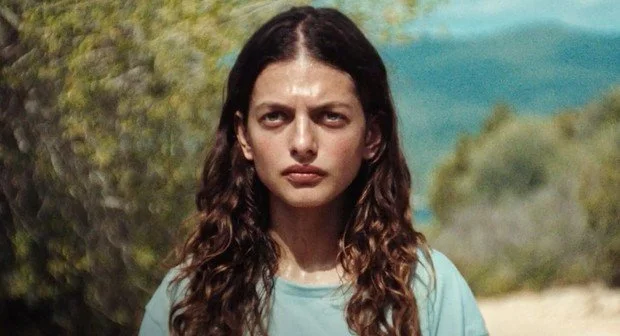 A woman with long brown curly hair stands outdoors with trees and mountains in the background, wearing a light blue shirt.