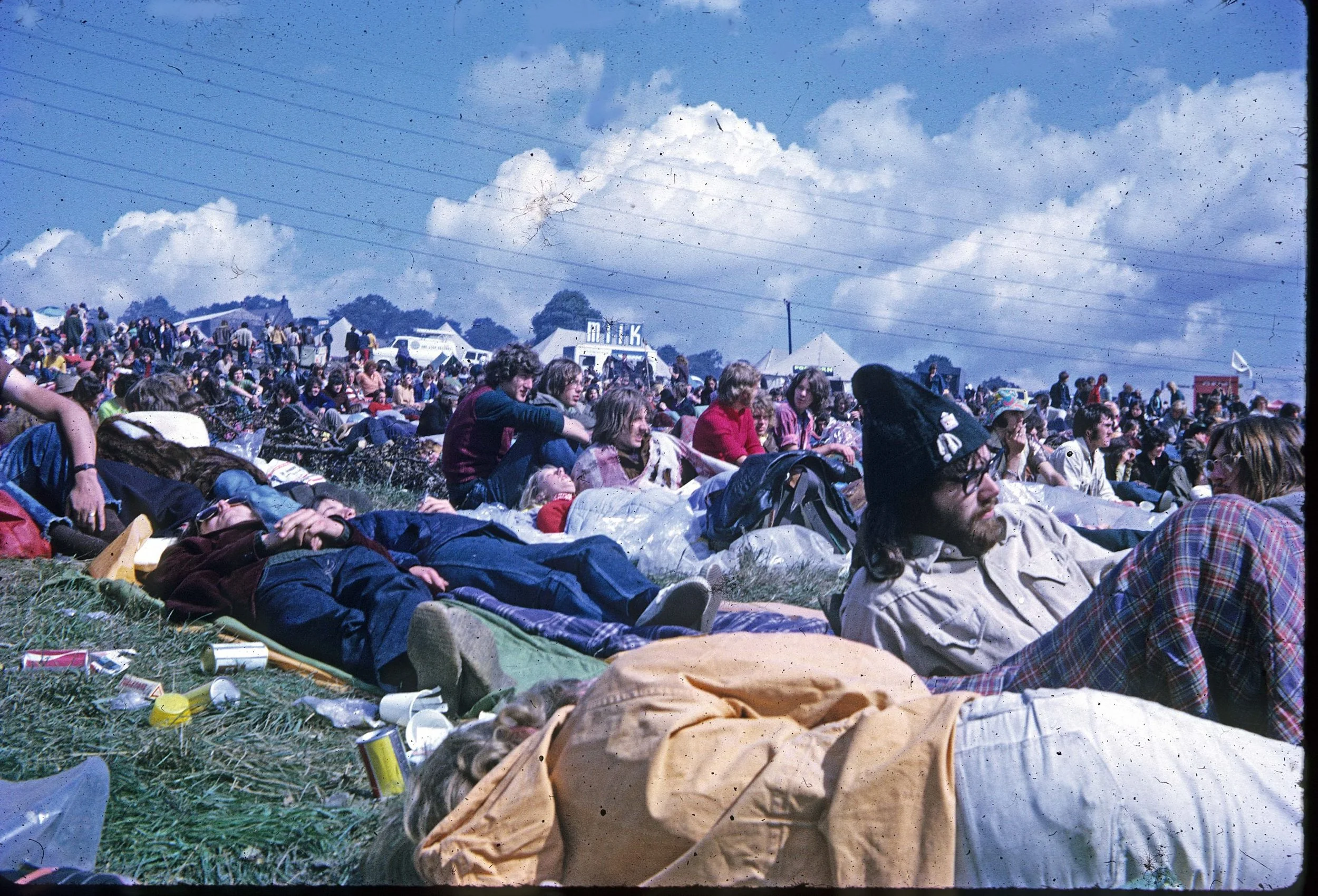 Large crowd of people sitting and lying on the grass at an outdoor music festival or event under a partly cloudy sky, with tents and vehicles in the background.