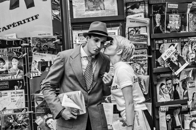 A young man in a suit and hat is holding a newspaper, while a young woman leans in to whisper in his ear in a magazine or bookstore, surrounded by magazines and books.