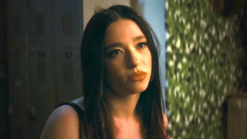 A woman with long dark hair and lipstick looking thoughtfully into the distance in an indoor setting with lockers and greenery.