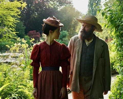 A woman and a man dressed in vintage clothing walking in a lush garden or park with green trees and plants.