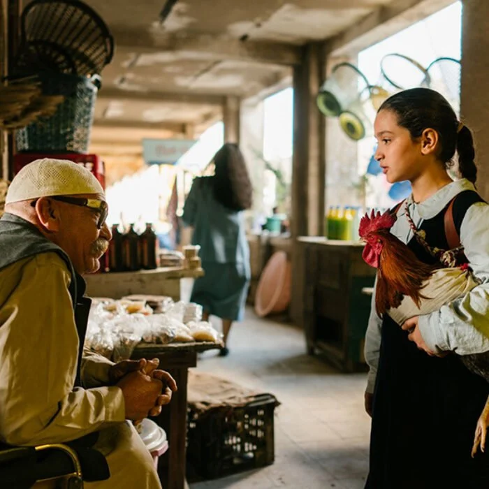 A woman selling chickens at an outdoor market, talking to a man in a wheelchair. The market stall has bread and other goods, with a rustic wooden background and natural light.