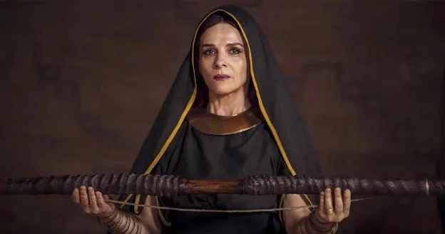 A woman dressed in black with a black head covering and gold accents, holding a staff horizontally in front of her, with a serious expression.