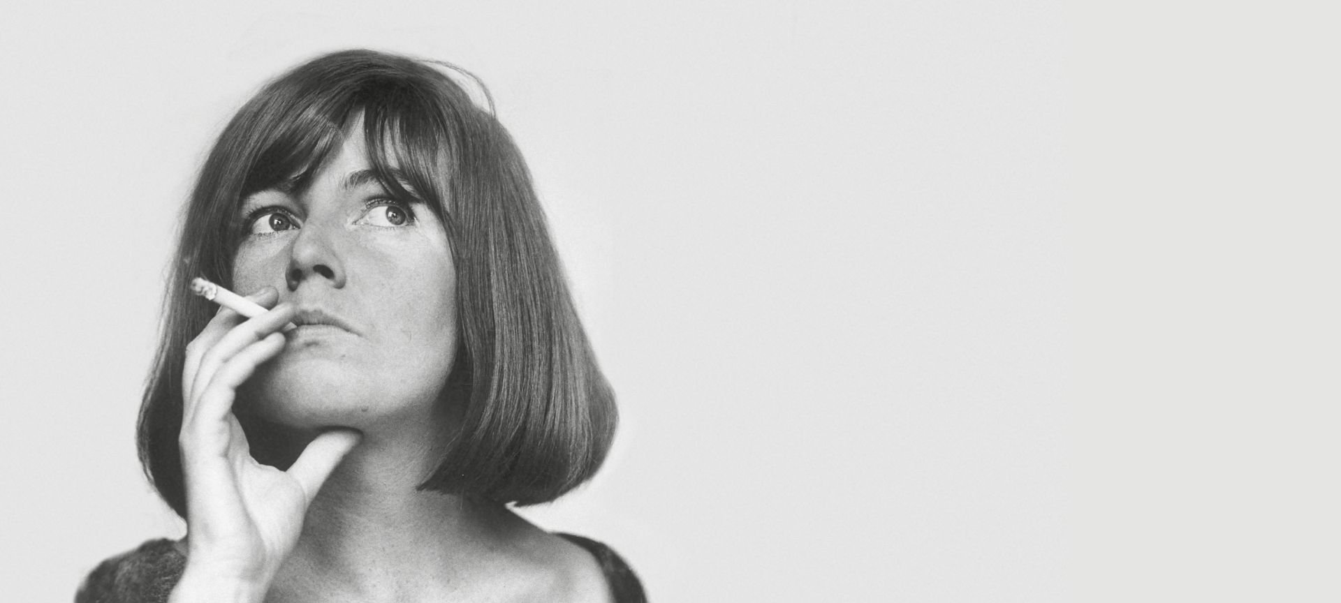 Black and white photo of a woman with a short dark hair, holding a cigarette near her mouth, looking upwards.