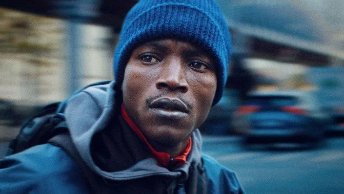 Close-up of a man wearing a blue beanie and a jacket, with a blurred urban background.