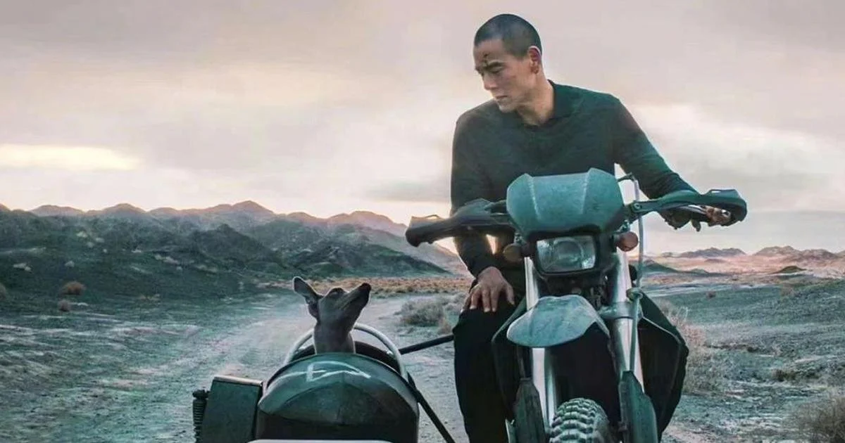 A man and a dog on an adventure motorcycle in the desert with mountains in the background.