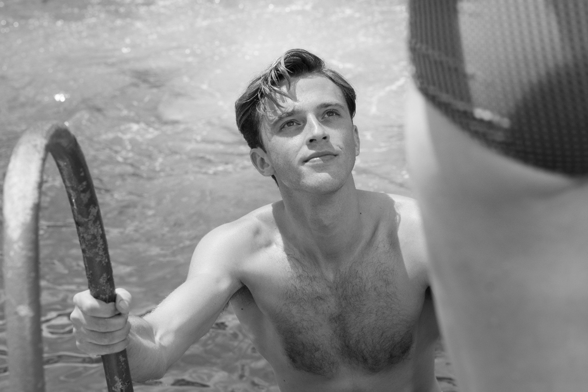 A black-and-white photo of a young man emerging from a swimming pool, holding a metal ladder, with water in the background, looking slightly upwards.