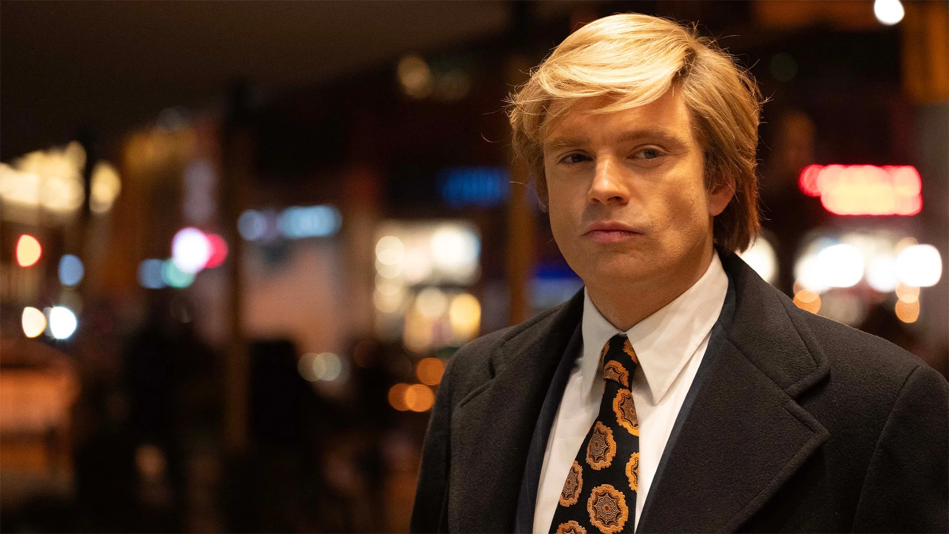 A man with blonde hair wearing a dark suit and patterned tie standing in a dimly lit bar or restaurant, with blurred colorful lights in the background.
