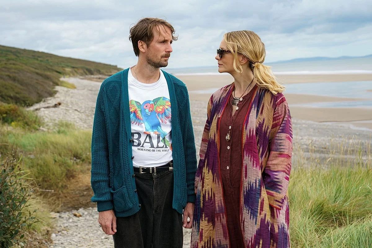 A man and a woman stand on a beach with grass and shrubbery, facing each other with serious expressions.