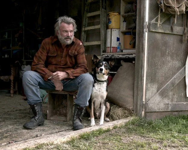 A bearded man in a brown flannel shirt and jeans sitting on a wooden stool outside a rustic barn, with a black and white dog sitting next to him. The scene is outdoors with grass in the foreground and barn tools and shelves in the background.