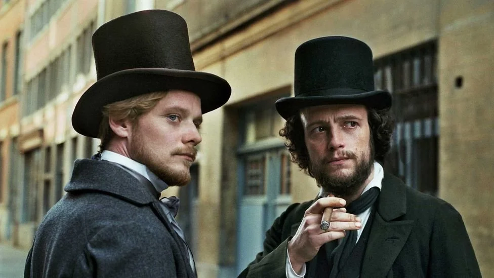Two men dressed in Victorian-era clothing, wearing tall black top hats, standing outdoors in a city street with old brick buildings.