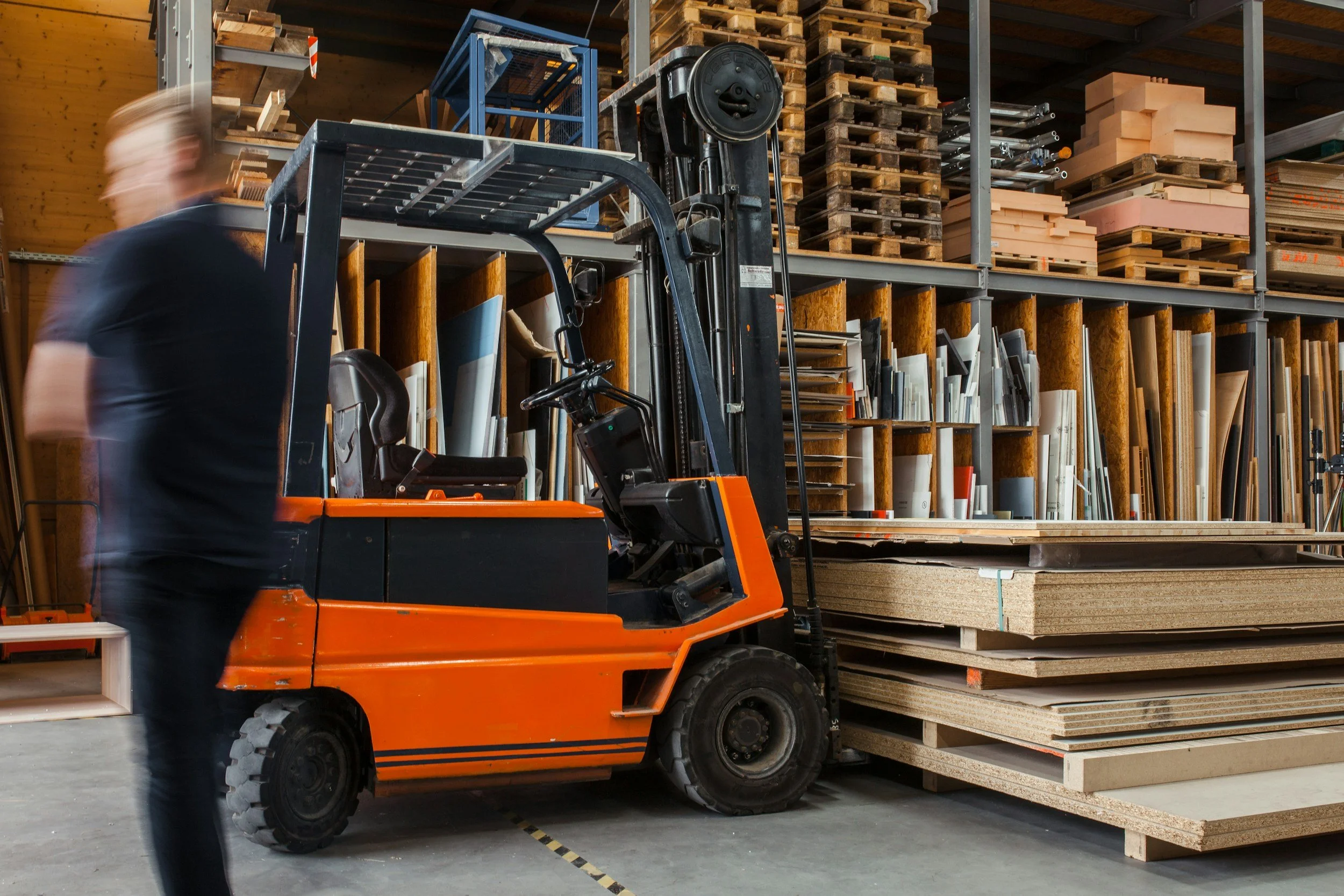 Blurred person walking past an orange forklift in a warehouse with stacks of plywood and shelving.