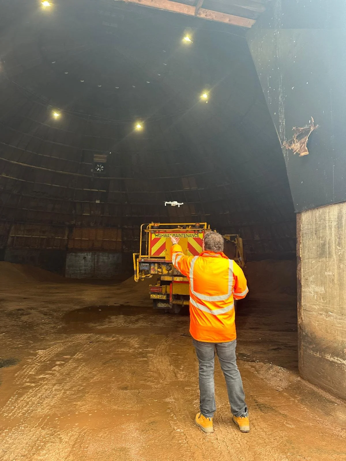 A worker in an orange high-visibility jacket and gray pants is inside a large underground tunnel, pointing at a maintenance vehicle. The tunnel has a curved, dark ceiling with lights and a drone flying near the ceiling. The ground is uneven and muddy.